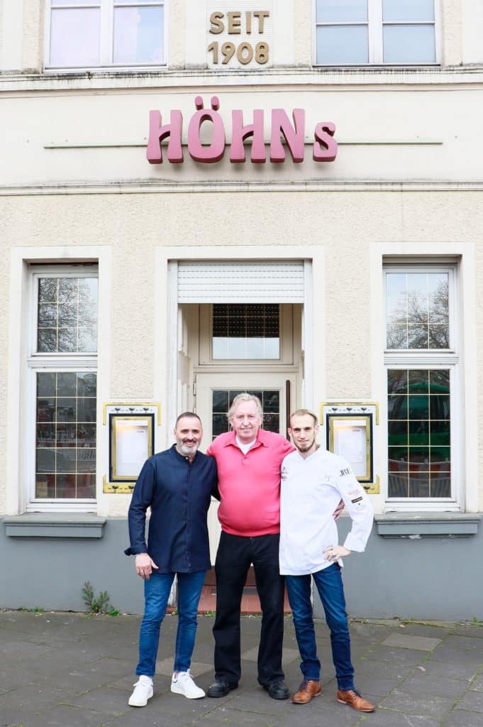 Luis Dias, Klaus Höhn und Cyril Becker stehen vor dem alten Höhns Gebäude in Köln mit dem Schild "Höhns" und "Seit 1908"