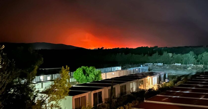 Das Foto zeigt Rheinenhäuser in einer Hotelanlage bei Nacht, mit den fernen Waldbränden, die den hügeligen Horizont Griechenlands erhellen.