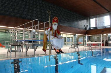 Das Foto zeigt eine Person in voller Nikolausverkleidung, die am Ende eines Sprungbretts in einer Schwimmhalle sitzt, die Beine lässig baumelnd. Der Nikolaus trägt eine rote und goldene Bischofsrobe, eine traditionelle Mitra auf dem Kopf und hält einen goldenen Bischofsstab, während er moderne schwarze Turnschuhe mit orangefarbenen Akzenten trägt.