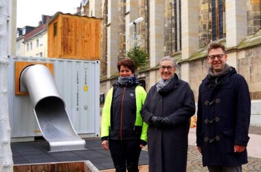 Das Foto zeigt drei Personen, die vor einem großen, metallischen Spielcontainer mit einer Rutsche stehen. Links steht Esther Weirauch, erkennbar an ihrer sportlichen Bekleidung und leuchtend gelben Jacke. In der Mitte steht Oberbürgermeisterin Sibylle Keupen, die eine stilvolle graue Jacke und eine schwarze Hose trägt, und rechts steht Kai Hennes, gekleidet in einen dunkelblauen Mantel mit traditionellen Knebelverschlüssen. Sie stehen auf einem gepflasterten Platz vor dem Container, der durch seine moderne Konstruktion in einem auffälligen Kontrast zu dem historischen Kirchenfenster im Hintergrund steht. Der Container selbst ist grau, mit einer hölzernen Oberseite und einer gebogenen silbernen Rutsche, die vom Inneren des Containers nach außen führt. Die Personen scheinen den neuen Spielbereich fröhlich einzuweihen.