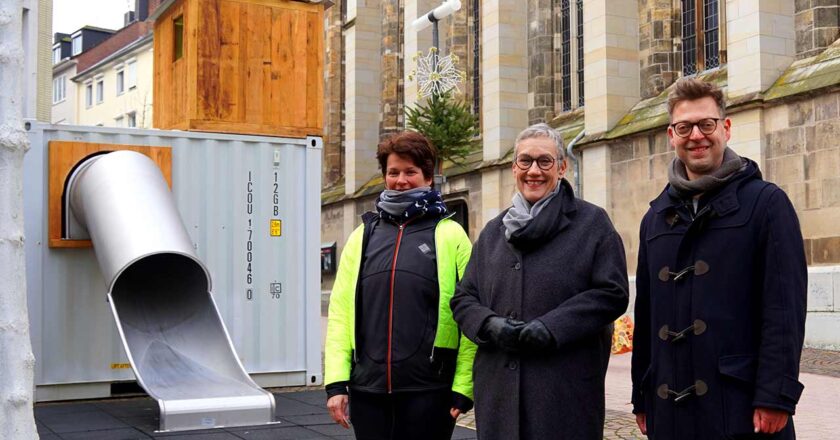 Das Foto zeigt drei Personen, die vor einem großen, metallischen Spielcontainer mit einer Rutsche stehen. Links steht Esther Weirauch, erkennbar an ihrer sportlichen Bekleidung und leuchtend gelben Jacke. In der Mitte steht Oberbürgermeisterin Sibylle Keupen, die eine stilvolle graue Jacke und eine schwarze Hose trägt, und rechts steht Kai Hennes, gekleidet in einen dunkelblauen Mantel mit traditionellen Knebelverschlüssen. Sie stehen auf einem gepflasterten Platz vor dem Container, der durch seine moderne Konstruktion in einem auffälligen Kontrast zu dem historischen Kirchenfenster im Hintergrund steht. Der Container selbst ist grau, mit einer hölzernen Oberseite und einer gebogenen silbernen Rutsche, die vom Inneren des Containers nach außen führt. Die Personen scheinen den neuen Spielbereich fröhlich einzuweihen.