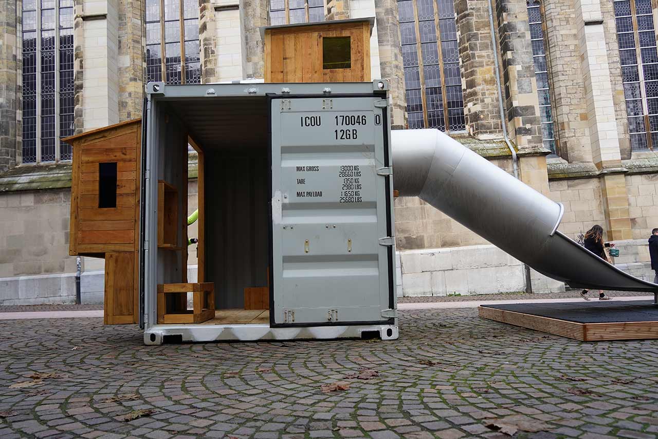 Das Foto zeigt einen grauen Schiffscontainer, der zu einem Spielmodul umfunktioniert wurde, mitten in einer Fußgängerzone. An der Seite des Containers ist eine große silberne Rutsche angebracht, die auf eine hölzerne Plattform führt. Auf dem Dach des Containers ist ein kleines Holzbüdchen mit einer Luke zu sehen. Der Container ist mit Kletterbalken ausgestattet, die an der Innenseite sichtbar sind. Im Hintergrund erkennt man das gotische Mauerwerk und die Fenster einer Kirche. Dieses innovative Spielmodul verbindet spielerisch funktionale Architektur mit urbanem Freiraum und bleibt für mindestens sechs Monate in der City Aachen