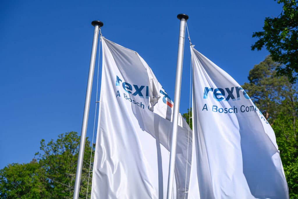 Zwei weiße Flaggen mit dem "Rexroth - A Bosch Company" Logo wehen im Wind vor einem klaren blauen Himmel und grünen Bäumen.