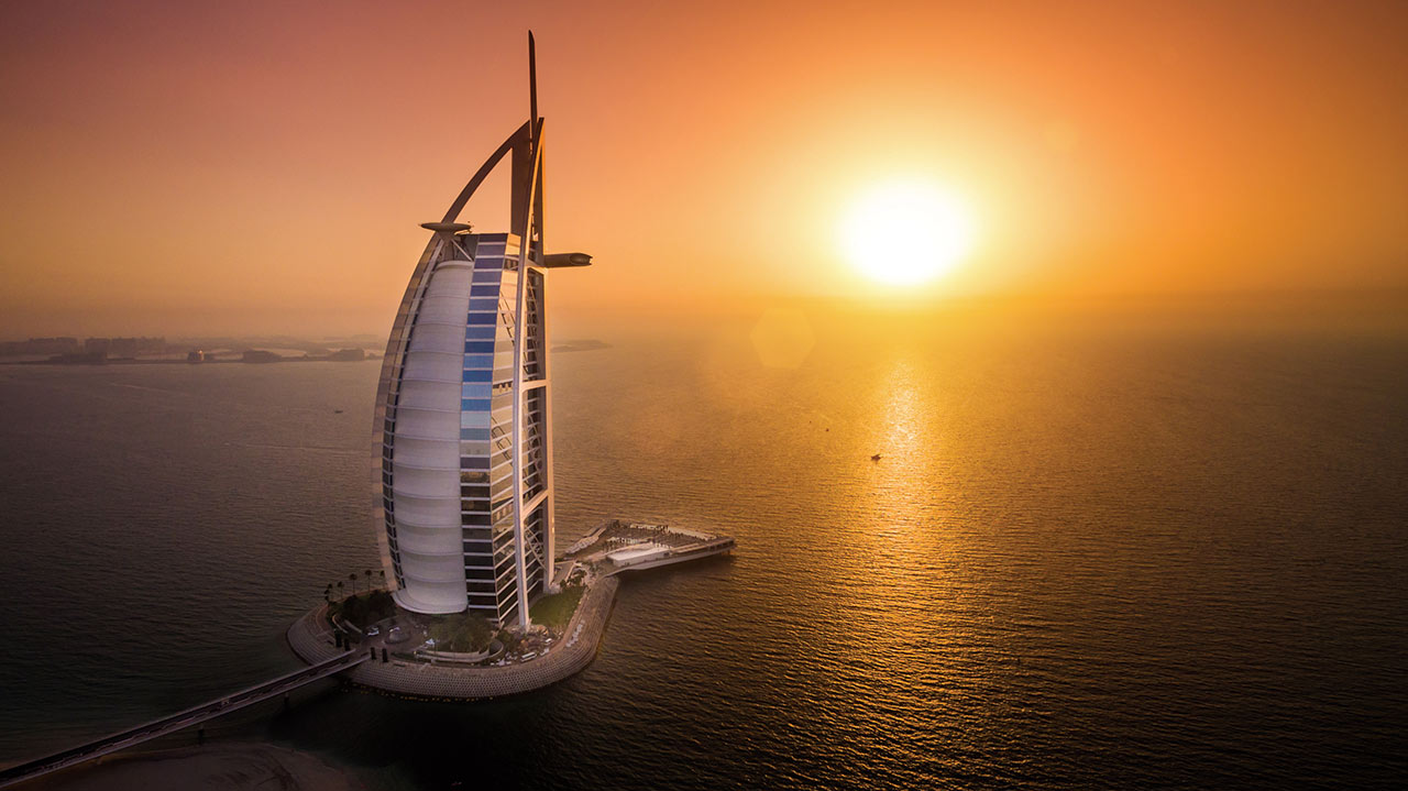 Das Foto zeigt den Burj Al Arab bei Sonnenuntergang, beleuchtet von den letzten Strahlen der untergehenden Sonne, umgeben vom Arabischen Golf.