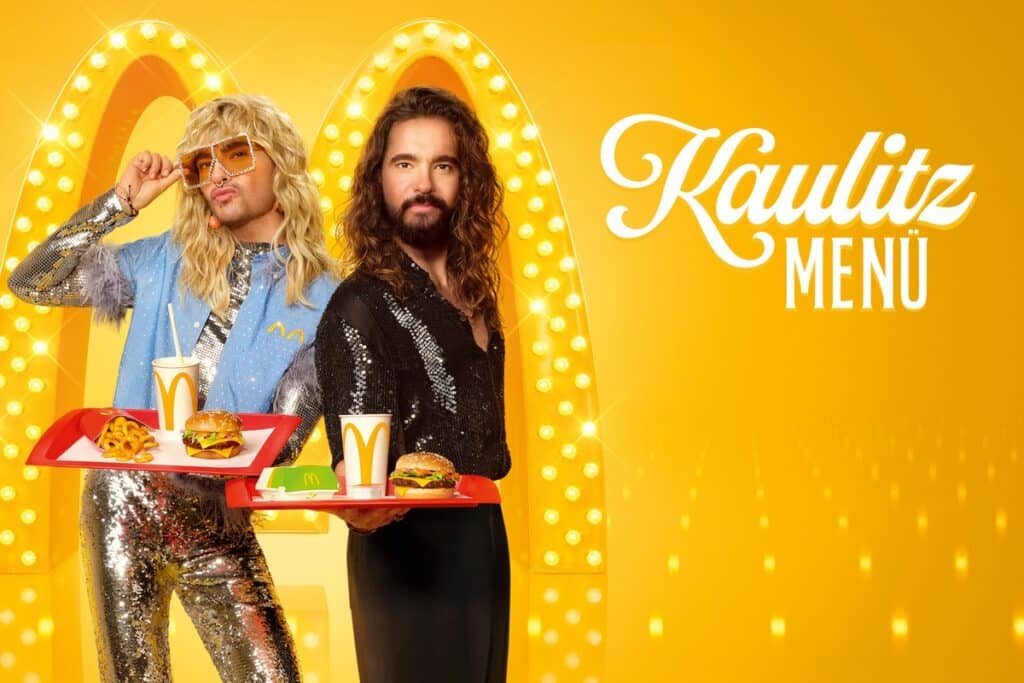 Bill und Tom Kaulitz präsentieren das Kaulitz Menü von McDonald’s, inklusive McPlant Burger, vor einem glamourösen, gelb beleuchteten Hintergrund.