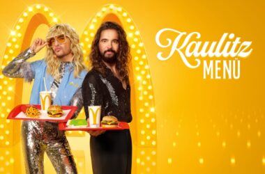 Bill und Tom Kaulitz präsentieren das Kaulitz Menü von McDonald’s, inklusive McPlant Burger, vor einem glamourösen, gelb beleuchteten Hintergrund.