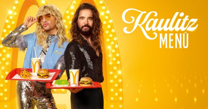 Bill und Tom Kaulitz präsentieren das Kaulitz Menü von McDonald’s, inklusive McPlant Burger, vor einem glamourösen, gelb beleuchteten Hintergrund.