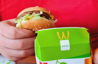 Eine Testperson von PlantPower-NoLimits.com trägt ein rotes T-Shirt mit dem Logo von PlantPower-NoLimits und hält einen McPlant Burger von McDonald's in der rechten Hand. In der linken Hand hält die Person die McPlant-Verpackung. Daneben ist eine teilweise sichtbare McPlant Nuggets-Box zu sehen. Die Verpackung von McDonald's zeigt ein veganes Symbol mit zwei Blättern und der Aufschrift „McPlant“.
