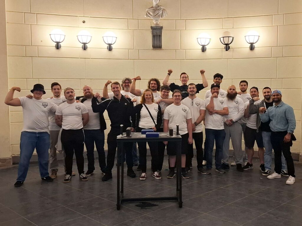 Gruppenfoto der Armwrestling Gruppe Aachen im Elisenbrunnen, mit Mitgliedern in weißer Kleidung, die freudig posieren. Michael Klassen und Roberto stehen lächelnd in der Mitte, umgeben von anderen Teammitgliedern, die ihre Armmuskeln zeigen.