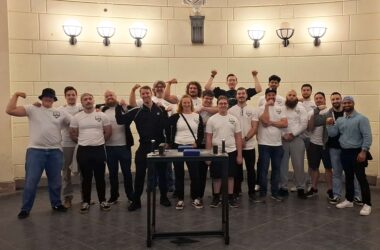 Gruppenfoto der Armwrestling Gruppe Aachen im Elisenbrunnen, mit Mitgliedern in weißer Kleidung, die freudig posieren. Michael Klassen und Roberto stehen lächelnd in der Mitte, umgeben von anderen Teammitgliedern, die ihre Armmuskeln zeigen.