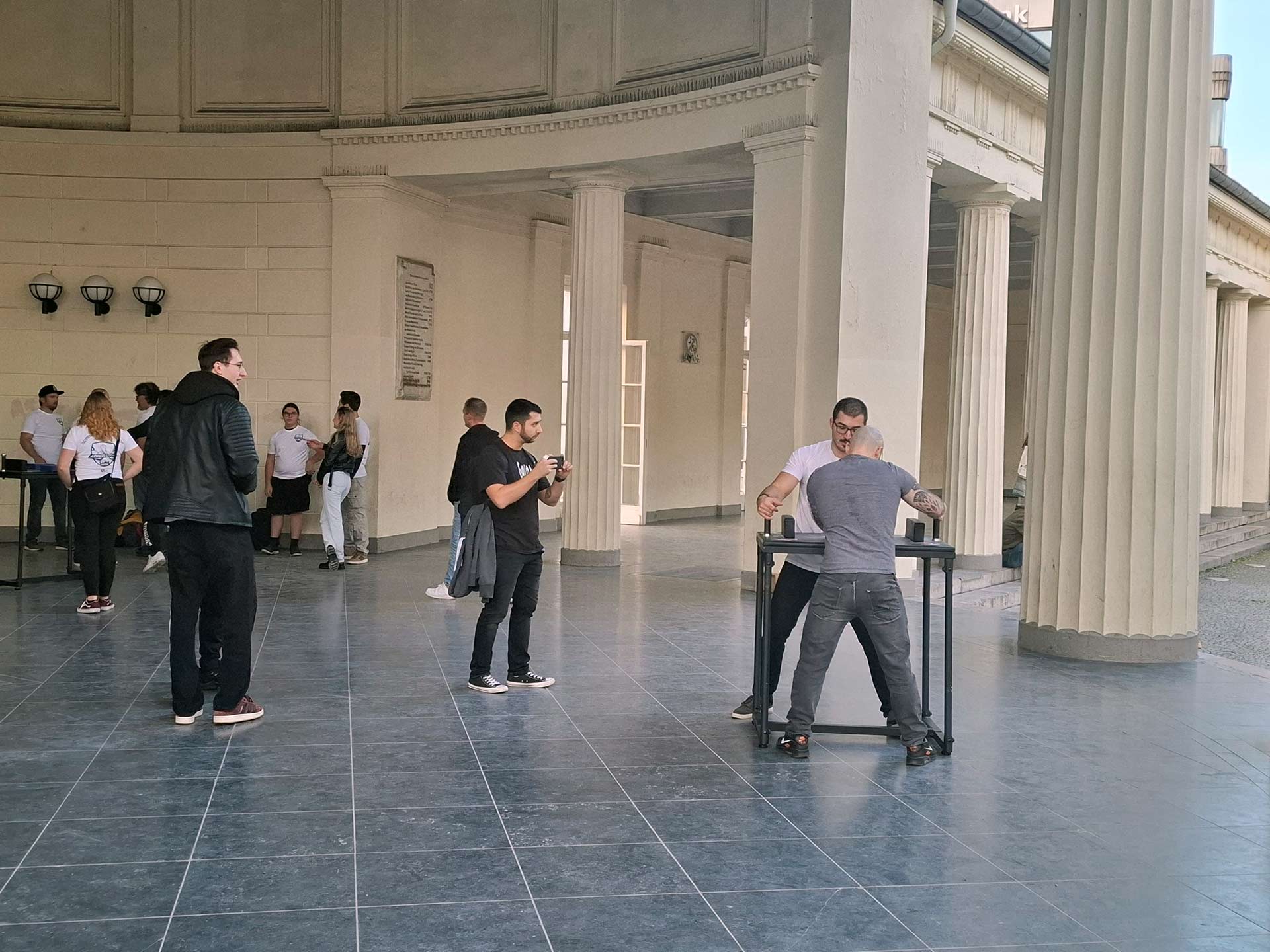 Ein friedliches Armwrestling-Match in Aachen am Elisenbrunnen, bei dem ein Profi im Vereins-T-Shirt und ein Zuschauer sich messen, unter den Augen einer kleinen Gruppe, inklusive eines Filmenden.
