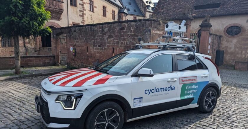 Ein weißes Vermessungsfahrzeug der CycloMedia Deutschland GmbH mit auf dem Dach montierter Kameratechnik steht auf einem gepflasterten Platz vor historischen Gebäuden.