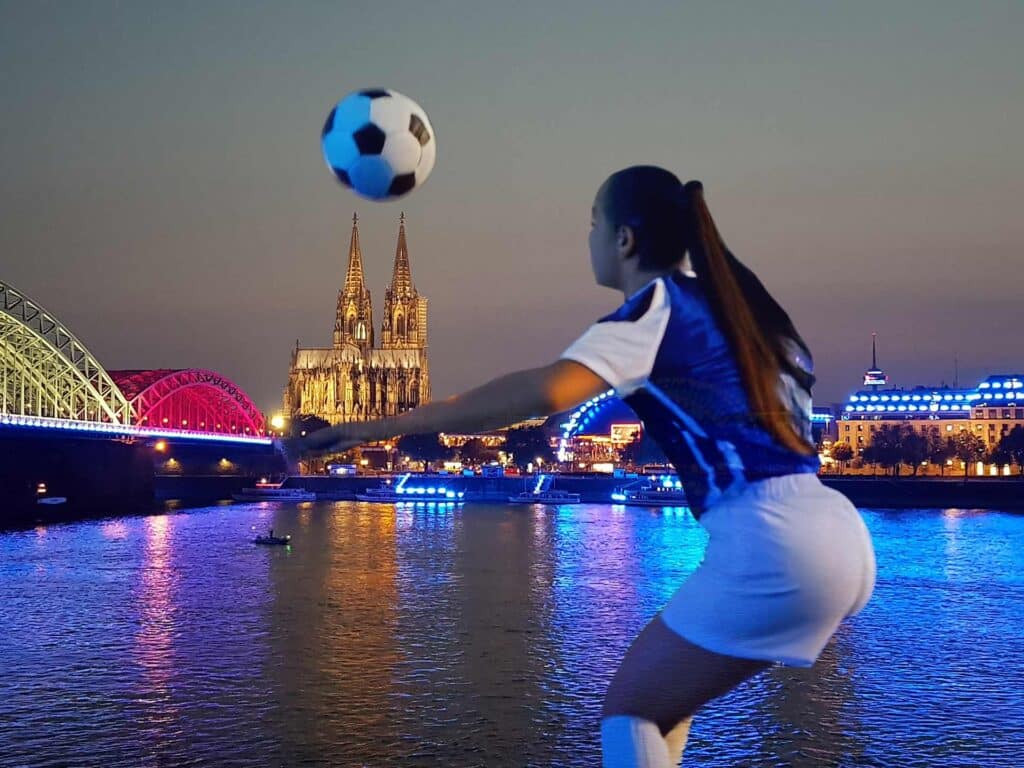 Eine Fußballspielerin in Aktion mit Blick auf den Kölner Dom und die Hohenzollernbrücke, die farbig beleuchtet über den Rhein erstrahlen.