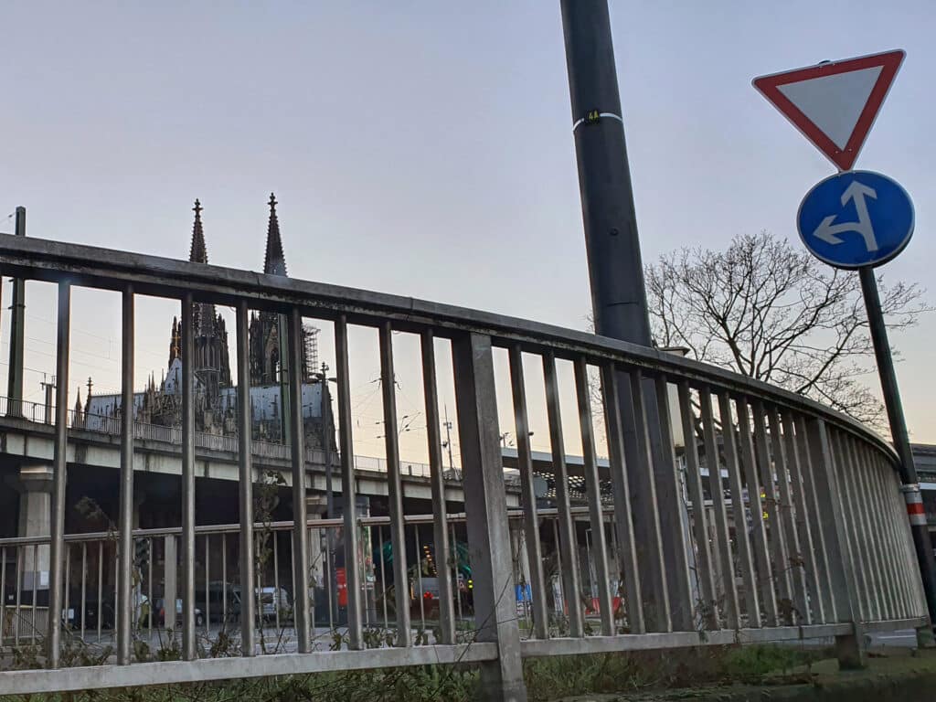 Blick durch ein Geländer auf den Kölner Dom, begleitet von Verkehrsschildern und urbaner Infrastruktur.