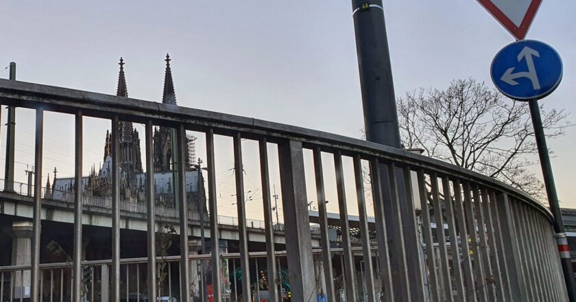 Blick durch ein Geländer auf den Kölner Dom, begleitet von Verkehrsschildern und urbaner Infrastruktur.