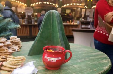 Ein dampfender roter Glühweinbecher mit der Aufschrift „Weihnachtsmarkt Bonn“ steht auf einem grünen Holztisch. Im Hintergrund sind verschwommen Weihnachtsmarktbuden und Lichter zu sehen.