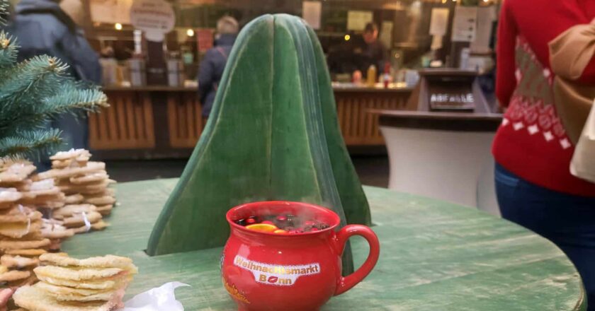 Ein dampfender roter Glühweinbecher mit der Aufschrift „Weihnachtsmarkt Bonn“ steht auf einem grünen Holztisch. Im Hintergrund sind verschwommen Weihnachtsmarktbuden und Lichter zu sehen.