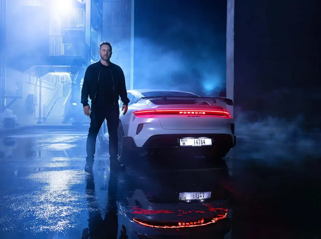 David Guetta steht neben einem weißen Porsche Taycan Turbo GT in einer stimmungsvollen, blau beleuchteten Kulisse mit reflektierendem Wasser am Boden.
