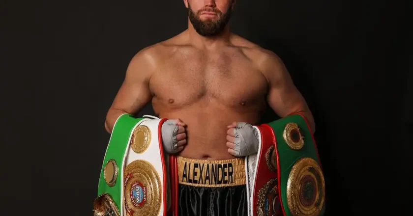 Profiboxer Alexander Frank steht mit mehreren Meisterschaftsgürteln vor schwarzem Hintergrund, oberkörperfrei und mit Bandagen an den Händen.