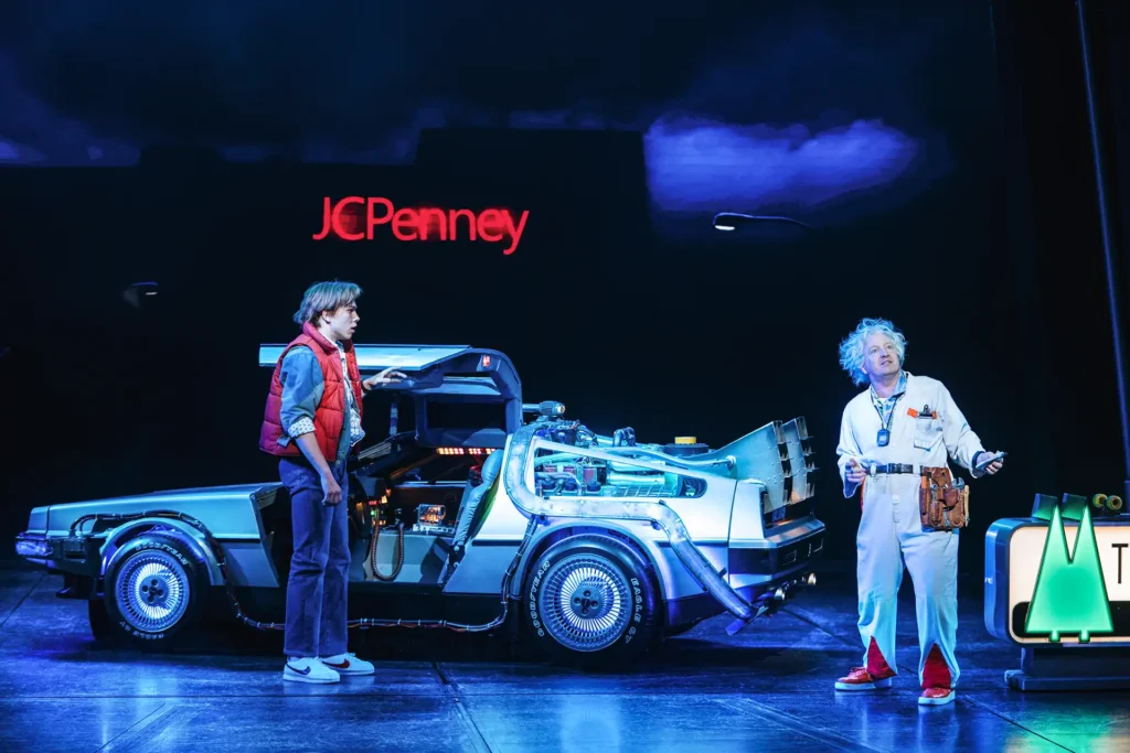 Zwei Schauspieler stehen auf einer Theaterbühne neben einem silbernen DeLorean mit geöffneten Flügeltüren. Der eine trägt eine rote Weste und Jeans, der andere einen weißen Kittel mit wildem, grauem Haar – eine Szene aus dem Musical „Zurück in die Zukunft“. Im Hintergrund leuchtet ein „JCPenney“-Schild.