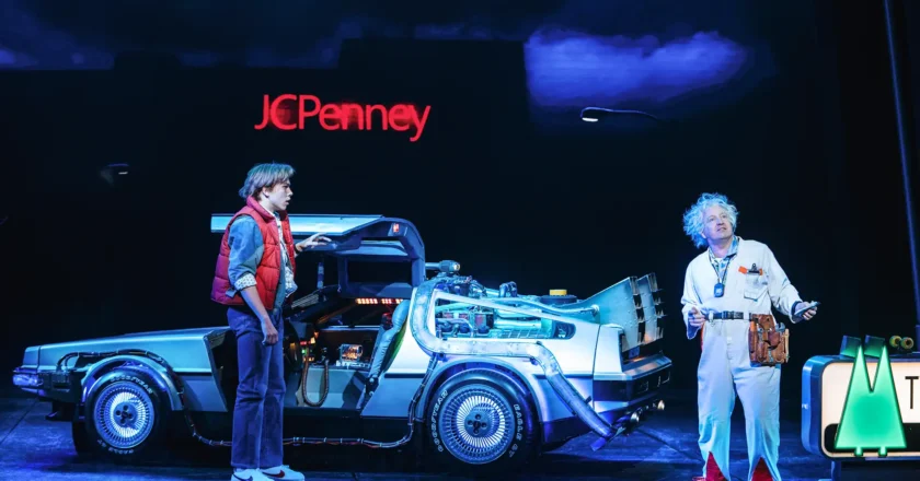 Zwei Schauspieler stehen auf einer Theaterbühne neben einem silbernen DeLorean mit geöffneten Flügeltüren. Der eine trägt eine rote Weste und Jeans, der andere einen weißen Kittel mit wildem, grauem Haar – eine Szene aus dem Musical „Zurück in die Zukunft“. Im Hintergrund leuchtet ein „JCPenney“-Schild.