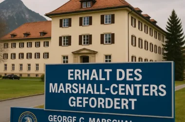 Das George C. Marshall Center in Garmisch-Partenkirchen ist bei Tageslicht zu sehen. Im Vordergrund steht ein großes blaues Schild mit der Aufschrift „ERHALT DES MARSHALL-CENTERS GEFORDERT“. Im Hintergrund das historische Hauptgebäude mit rotem Dach und Bergkulisse.