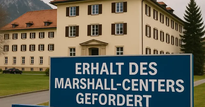Das George C. Marshall Center in Garmisch-Partenkirchen ist bei Tageslicht zu sehen. Im Vordergrund steht ein großes blaues Schild mit der Aufschrift „ERHALT DES MARSHALL-CENTERS GEFORDERT“. Im Hintergrund das historische Hauptgebäude mit rotem Dach und Bergkulisse.