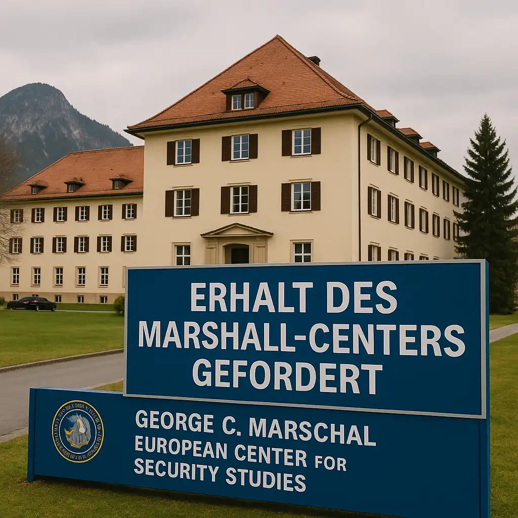 Das George C. Marshall Center in Garmisch-Partenkirchen ist bei Tageslicht zu sehen. Im Vordergrund steht ein großes blaues Schild mit der Aufschrift „ERHALT DES MARSHALL-CENTERS GEFORDERT“. Im Hintergrund das historische Hauptgebäude mit rotem Dach und Bergkulisse.