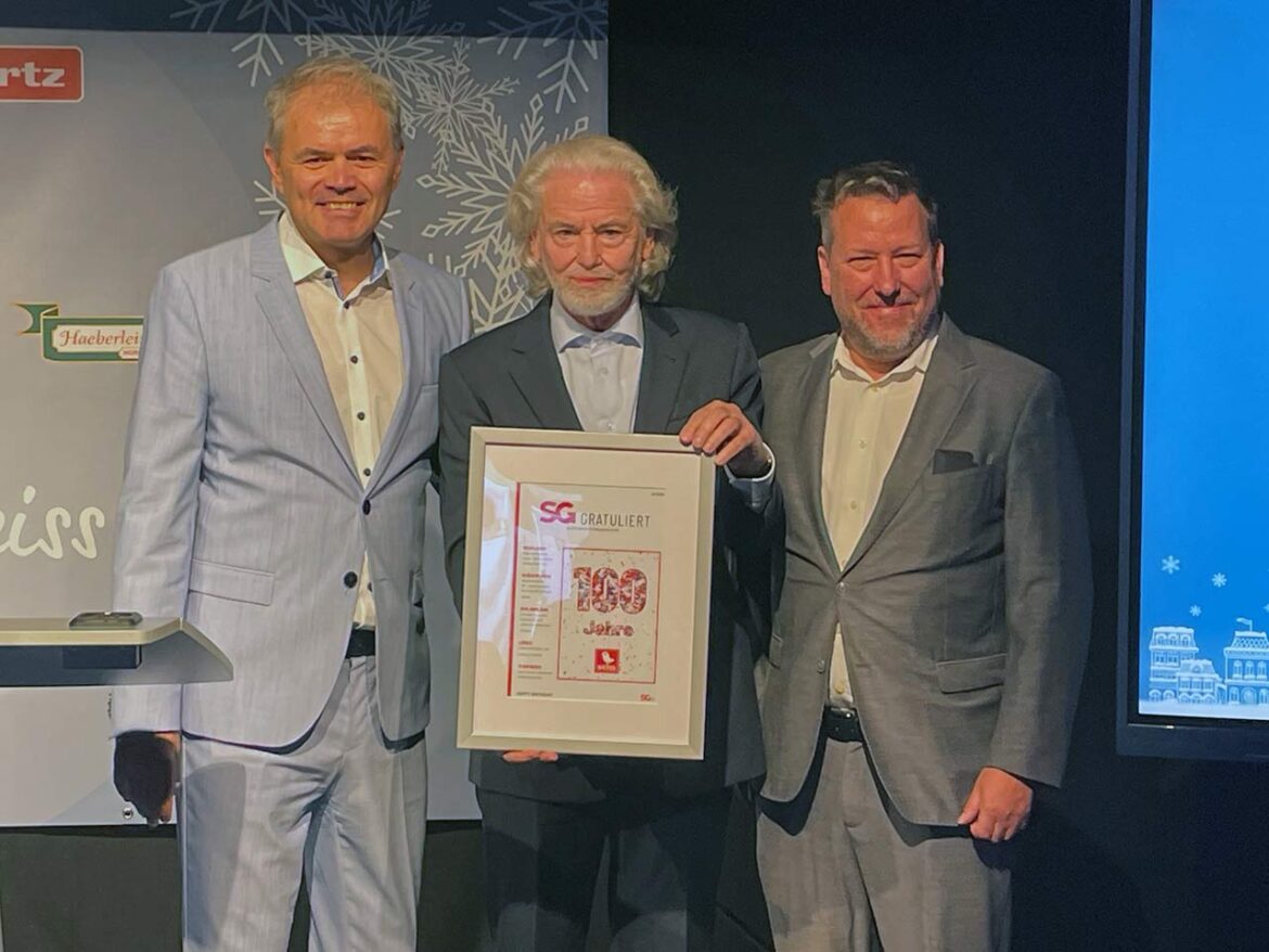 Drei Männer in Anzügen auf einer Bühne; die mittlere Person hält eine gerahmte Urkunde. Laut Angabe: links Joachim Eckert, Mitte Dr. Hermann Bühlbecker, rechts Oliver Peik.