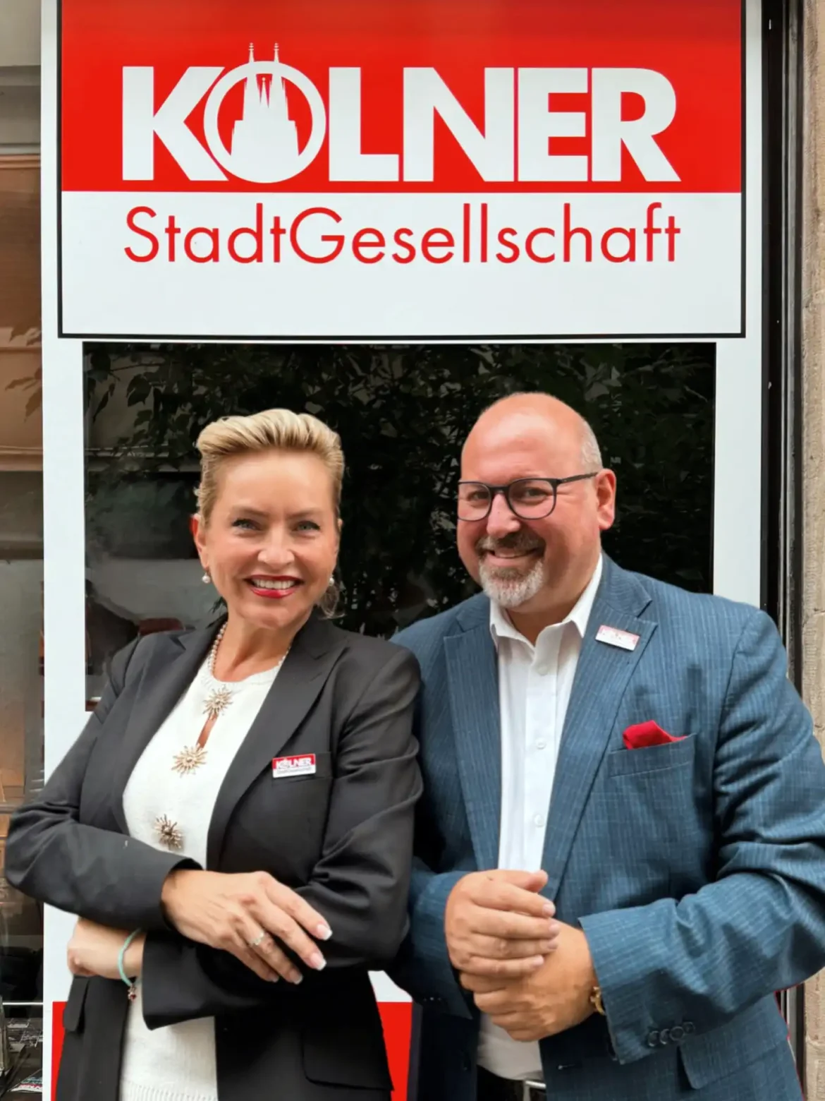 Links steht Harriet Krüger und rechts steht OP Kandidat Roberto Campione vor dem Kölnder StadtGesellschaft (KSG) - Logo, beide lächelnd.