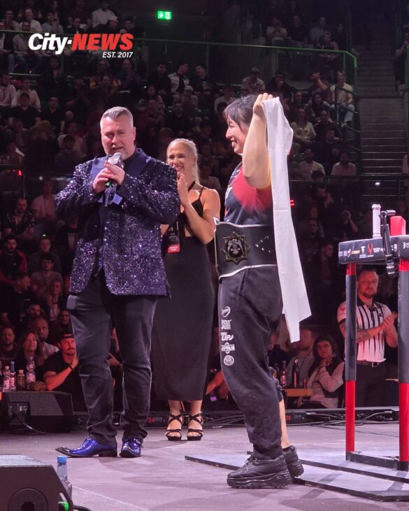 Ayane Takenaka stands on the East vs West 23 stage after defeating Melek Sahin 4-0 to win the Right Arm Featherweight World Title, holding the Japanese flag as host Neil Pickup speaks beside her and Sarah Bäckman stands behind.