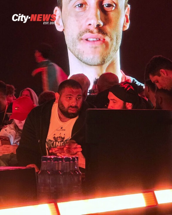 Levan Saginashvili watches the event floor from ringside while his enlarged image dominates the background screen, creating a striking double portrait of presence and status.